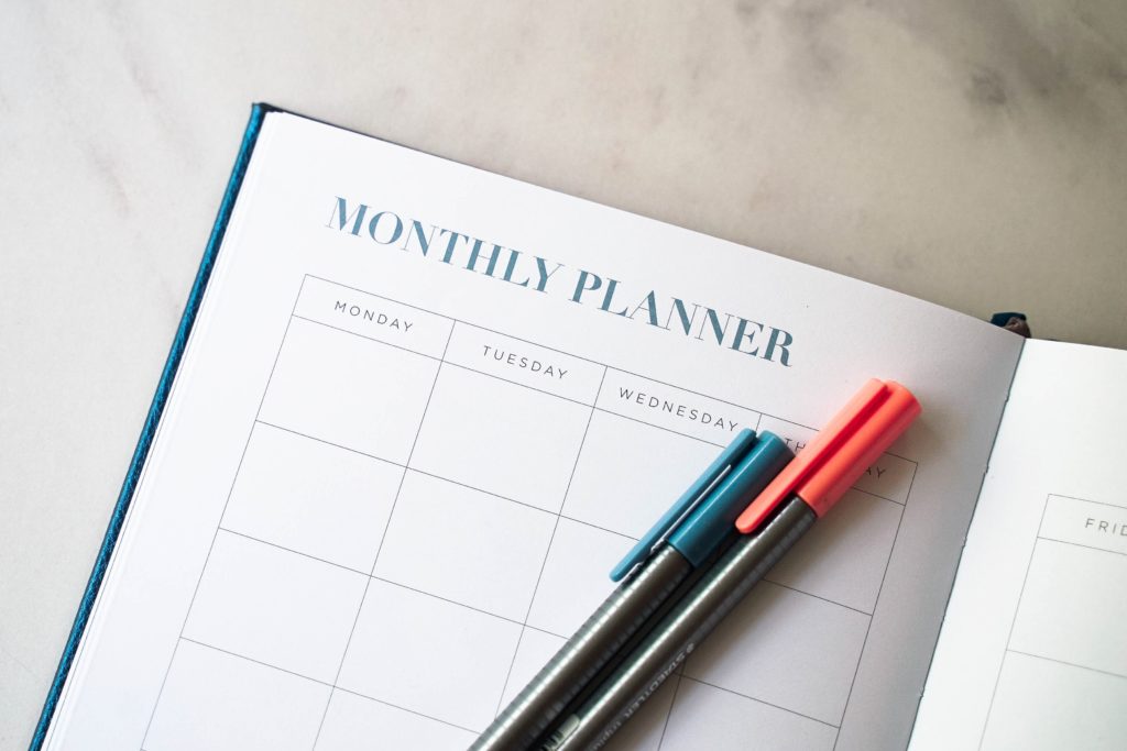 This open monthly planner has spaces for each day and green- and red-capped pens lying on it.