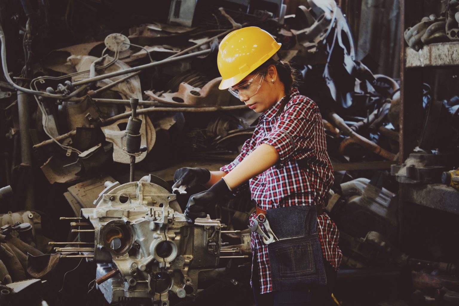 A woman wearing a yellow hard hat, goggles, and black gloves performs maintenance on an engine.